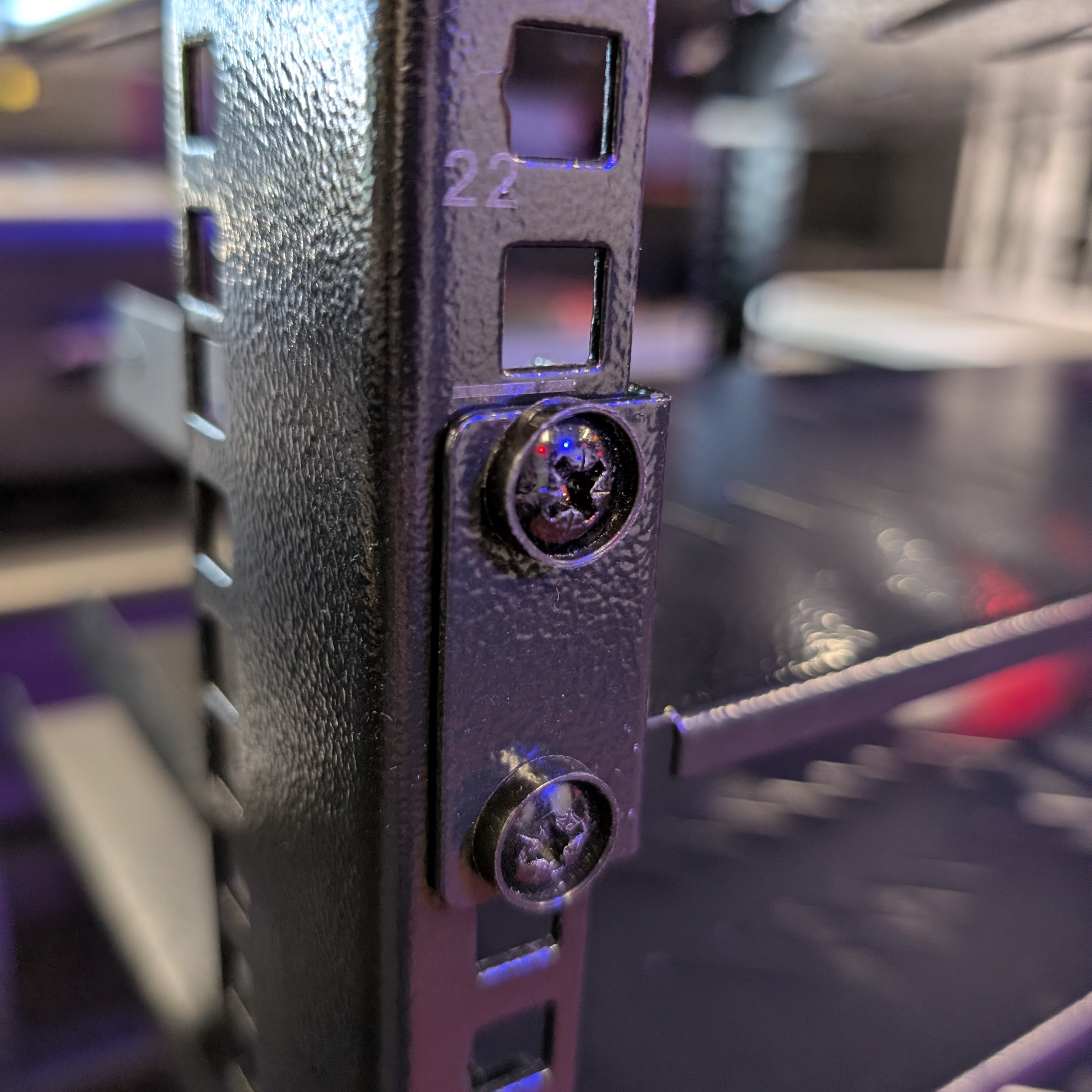 Close-up of a black metal server rack mounting bracket secured with two screws, showing the textured steel surface and depth slot details.
